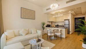 A modern living room with a sofa, a coffee table, and a dining area with chairs and a kitchen in the background. - Manzil -1BR w Pool nr Dubai Sports City  (Dubai)