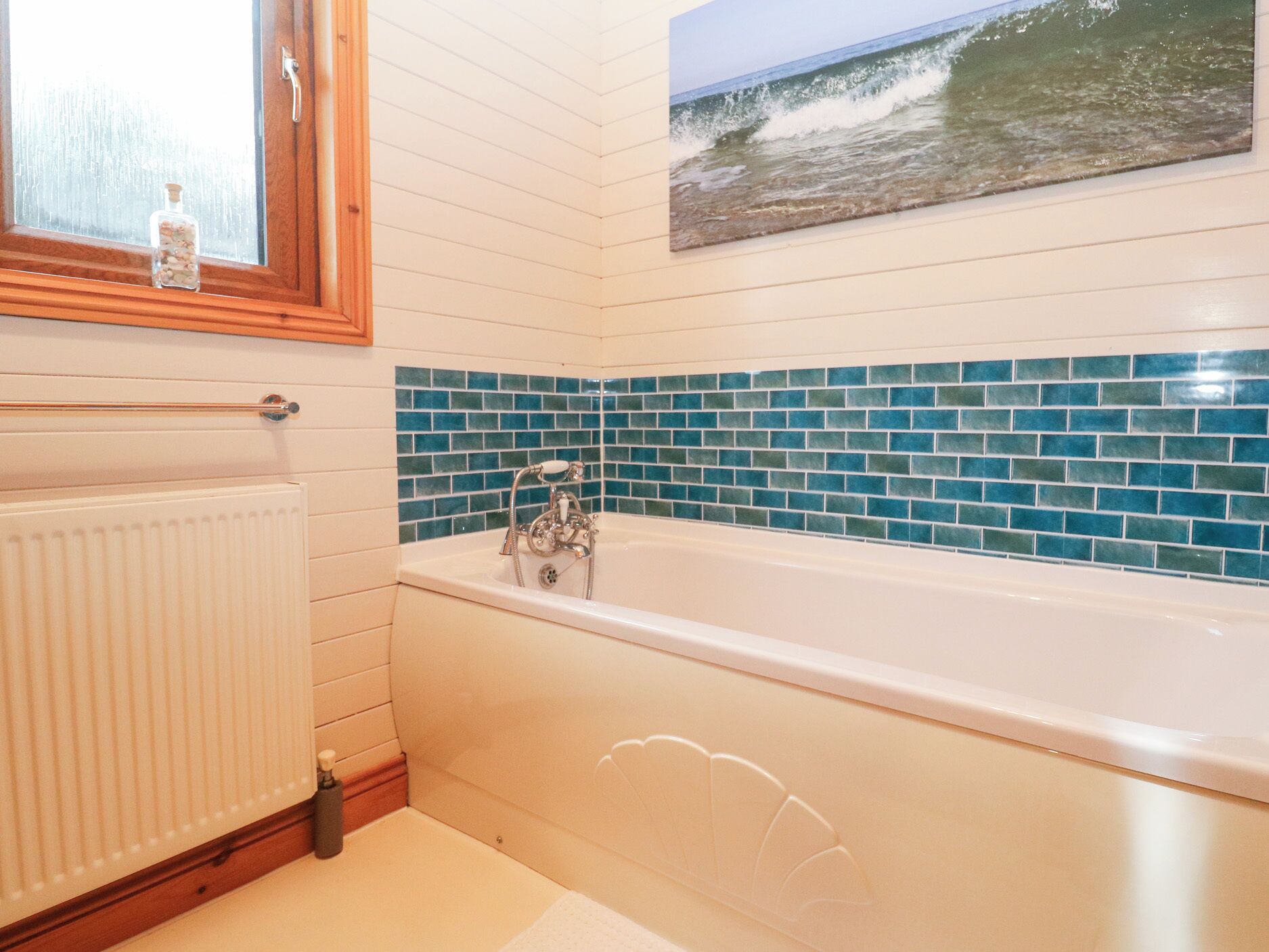 Un baño con una bañera blanca, paredes cubiertas de azulejos azules, un marco de ventana de madera y una imagen del océano.
