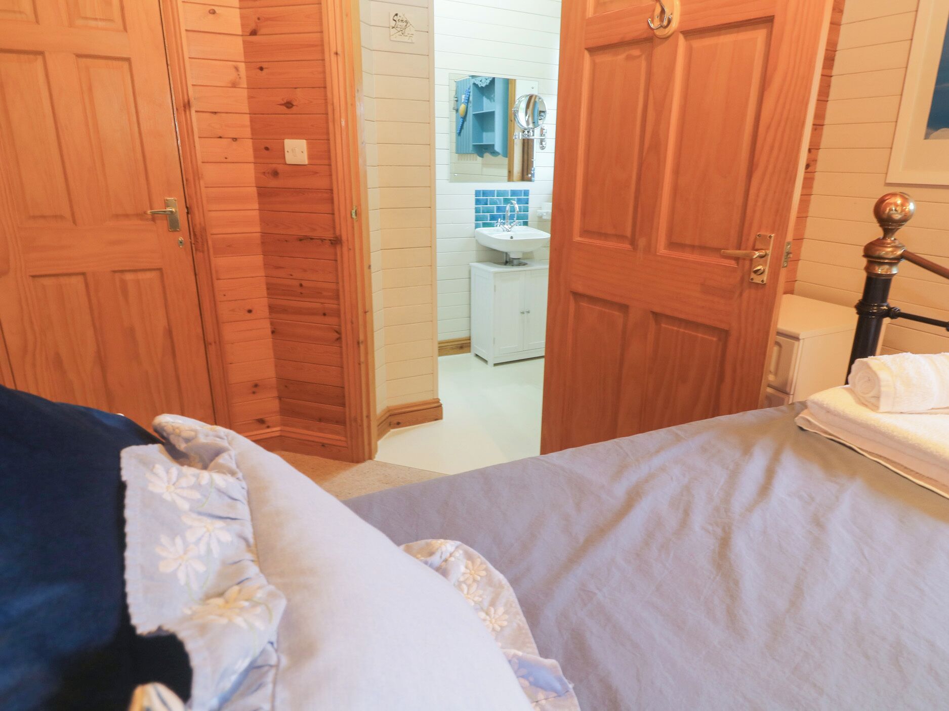 Een slaapkamer met een houten deur, een bed met een deken in blauw en wit, en een witte nachtkastje met een handdoek.