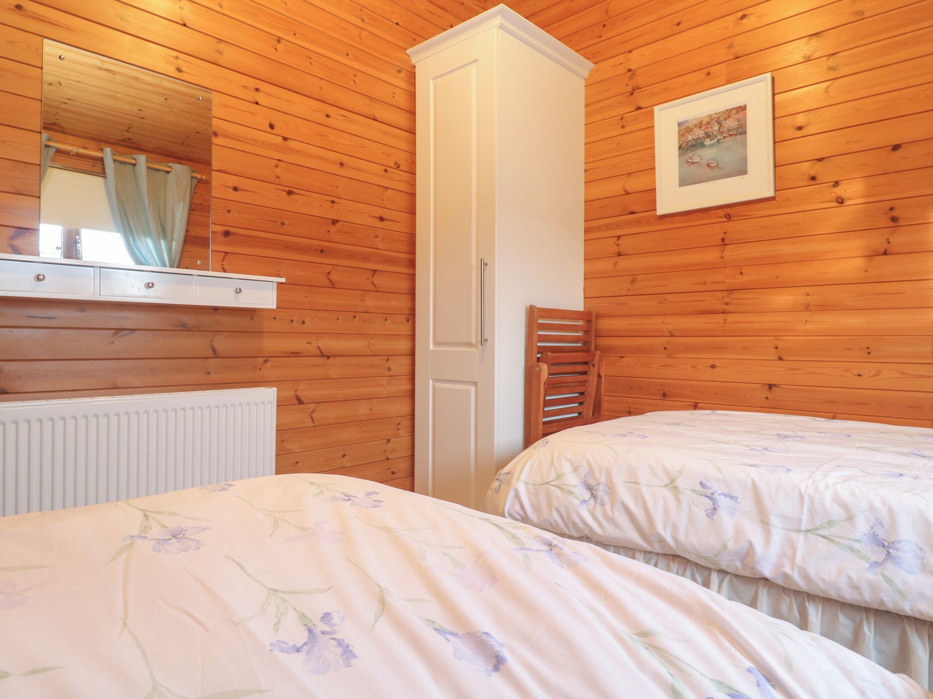 A bedroom with wooden panel walls, a white wardrobe, two beds with floral bedding, a radiator, and a framed picture on the wall.