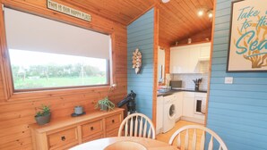 A cozy cabin interior with a dining table, chairs, a window with a view of the outdoors, a kitchen area with a washing machine, and a sign that reads "THIS IS OUR HAPPY PLACE". - Lamorna (Penzance)