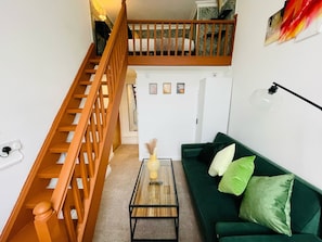 A staircase leading to an upper level, a green sofa with cushions, a small table with a vase, and a wall-mounted lamp.