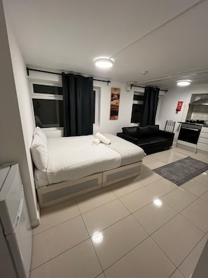 A modern hotel room with a bed, a sofa, a kitchenette, and a bathroom.