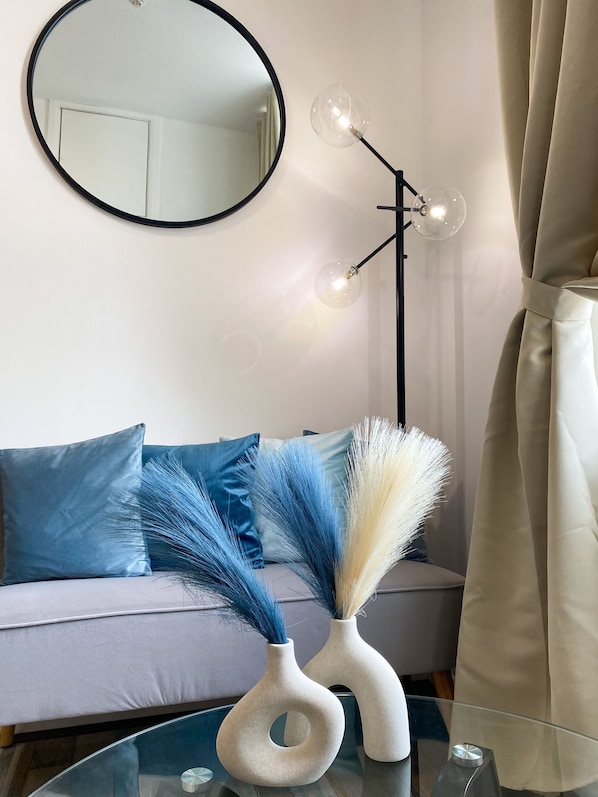 A room with a round mirror, a modern floor lamp, a sofa with blue and white cushions, and two vases with decorative grasses.
