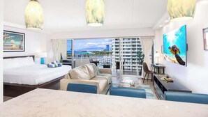 A hotel room with a bed, a sofa, a dining table, and a TV. There is a large window with a view of buildings and a marina. - Ocean & marina view w/ steps to Waikiki Beach (Honolulu)
