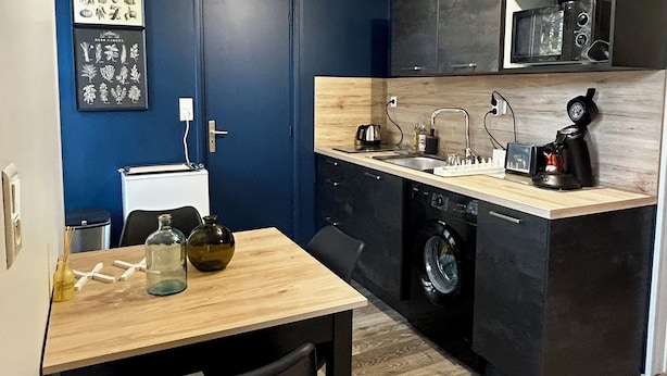 Cozinha compacta com micro-ondas, pia e máquina de lavar. Há uma mesa de jantar com cadeiras e um pequeno armário com um micro-ondas em cima.