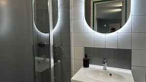 Uma casa de banho moderna com cabine de duche em vidro, lavatório branco e espelho grande com iluminação circular.