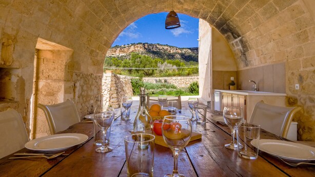 A dining table set with plates, glasses, and a fruit basket, with a view of a mountainous landscape through an arched stone structure. - Holiday Home 'Son Pons' with Mountain Views, Wi-Fi & Air Conditioning (Llucmajor)
