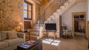 A stone-walled room with a staircase, a sofa, a television on a stand, and a rocking chair. - Holiday Home 'Son Pons' with Mountain Views, Wi-Fi & Air Conditioning (Llucmajor)