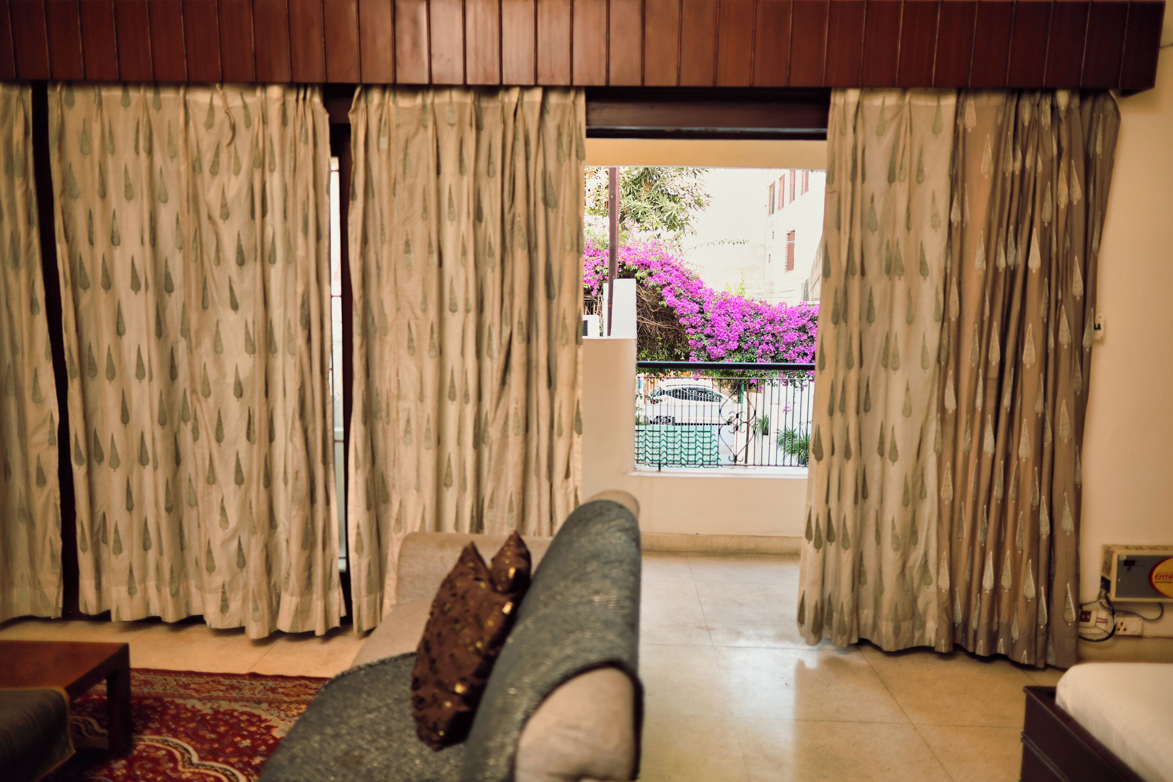 Una habitación con sofá, una mesita y una cama. Hay cortinas en las ventanas y se ve un balcón con flores rosas.