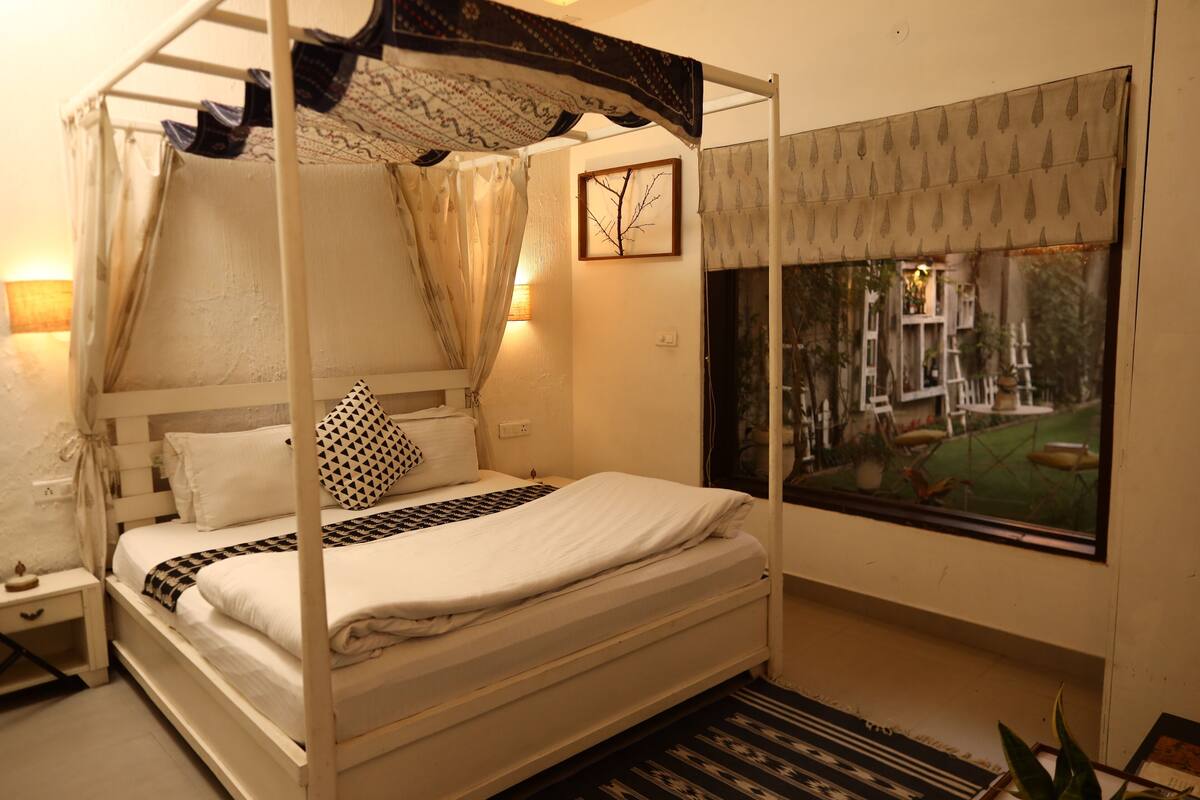 A four-poster bed with a canopy, a bedside table with a lamp, a framed picture on the wall, and a window with a view of the outdoors.