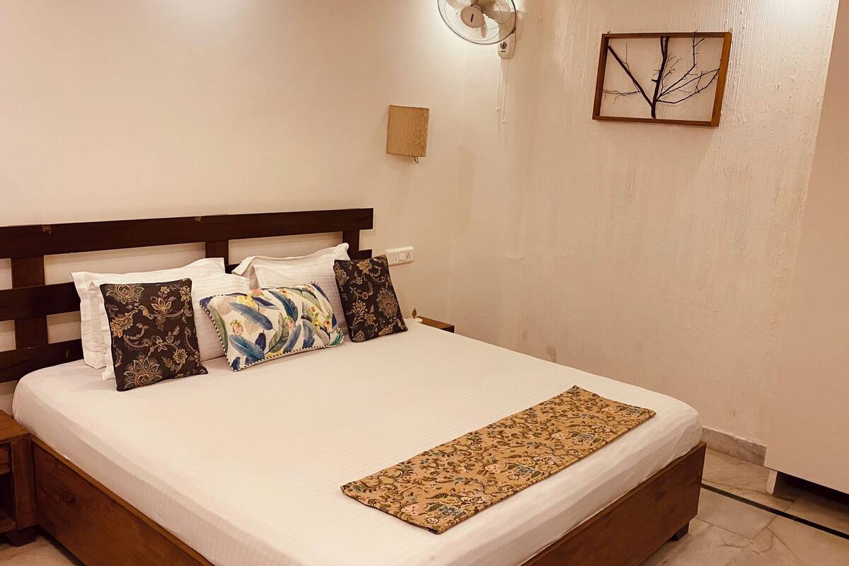 A neatly made bed with a wooden headboard, white bedding, and decorative pillows. There is a bedside table with a lamp and a wall-mounted artwork above the bed.