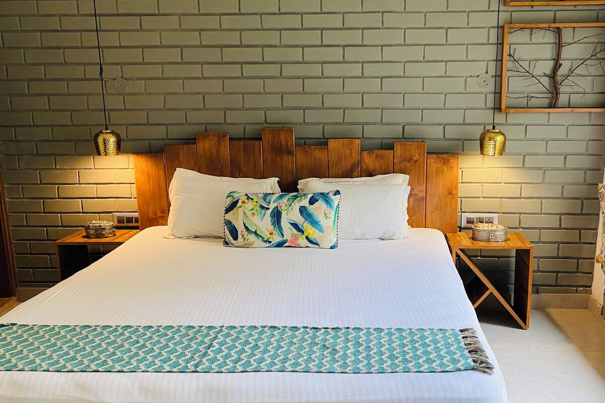 A neatly made bed with white linens, a patterned throw, and a wooden headboard. There are two bedside tables with lamps, a decorative wall piece above the bed, and a bench with cushions to the right.