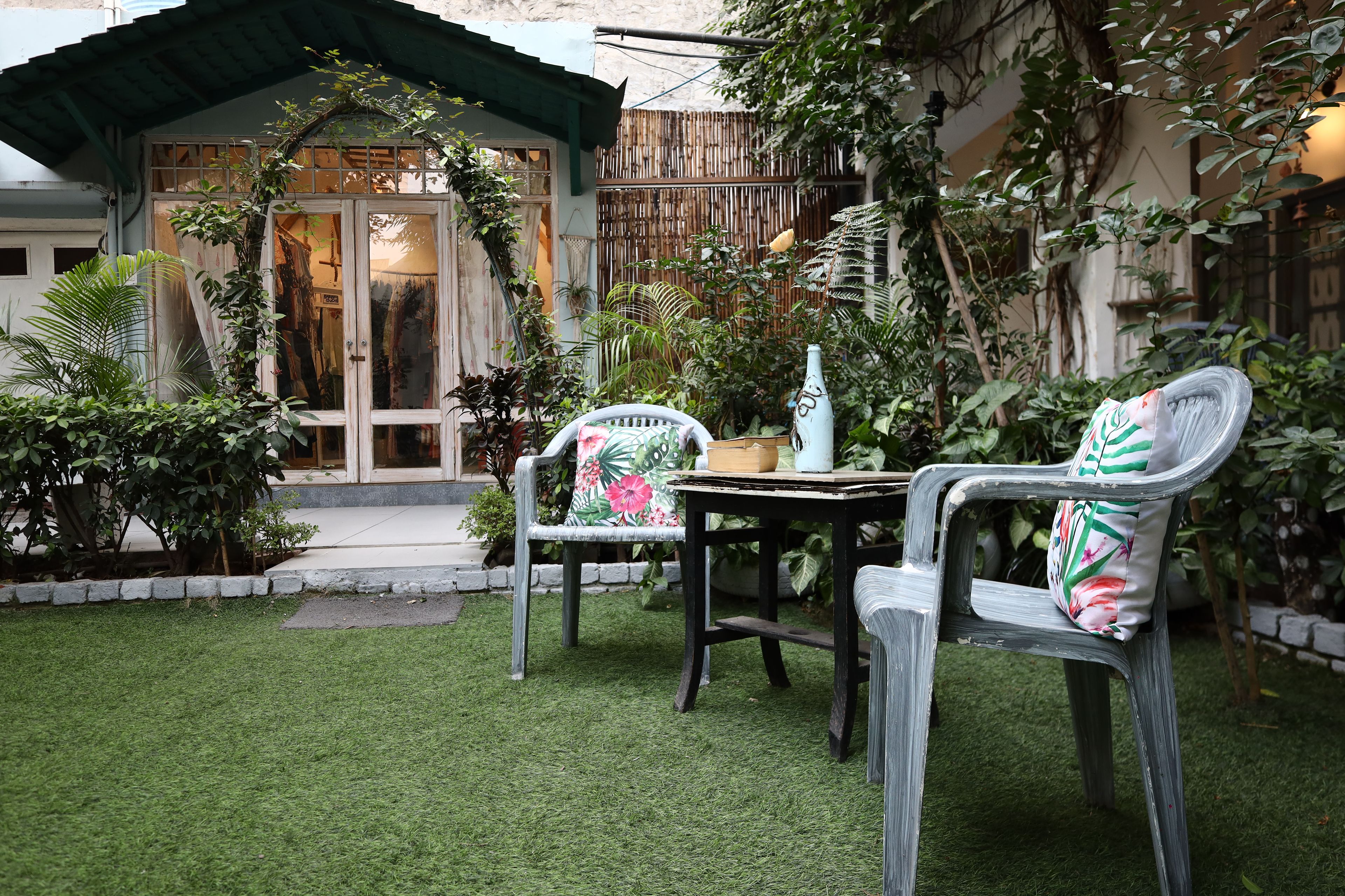 A patio with a table and two chairs, surrounded by lush greenery and a building with a green roof.