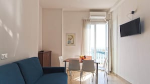 A living room with a sofa, a dining table with chairs, a TV mounted on the wall, and a balcony with a view. - Three-room seafront apartment with balcony (Alassio)