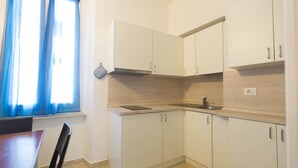 Una cuina petita amb armaris blancs, un rentapeus i una finestra amb cortines blaves.