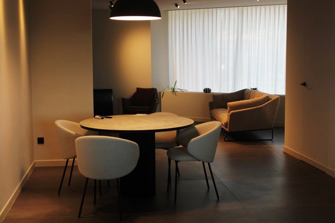 Un comedor moderno con una mesa redonda y cuatro sillas, un sofá y un amplio ventanal con cortinas.