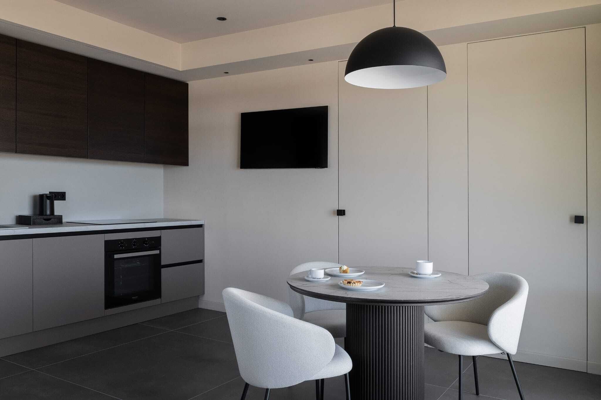 A modern kitchen with a dining area, featuring a round table, two chairs, a pendant light, and a flat-screen TV on the wall.