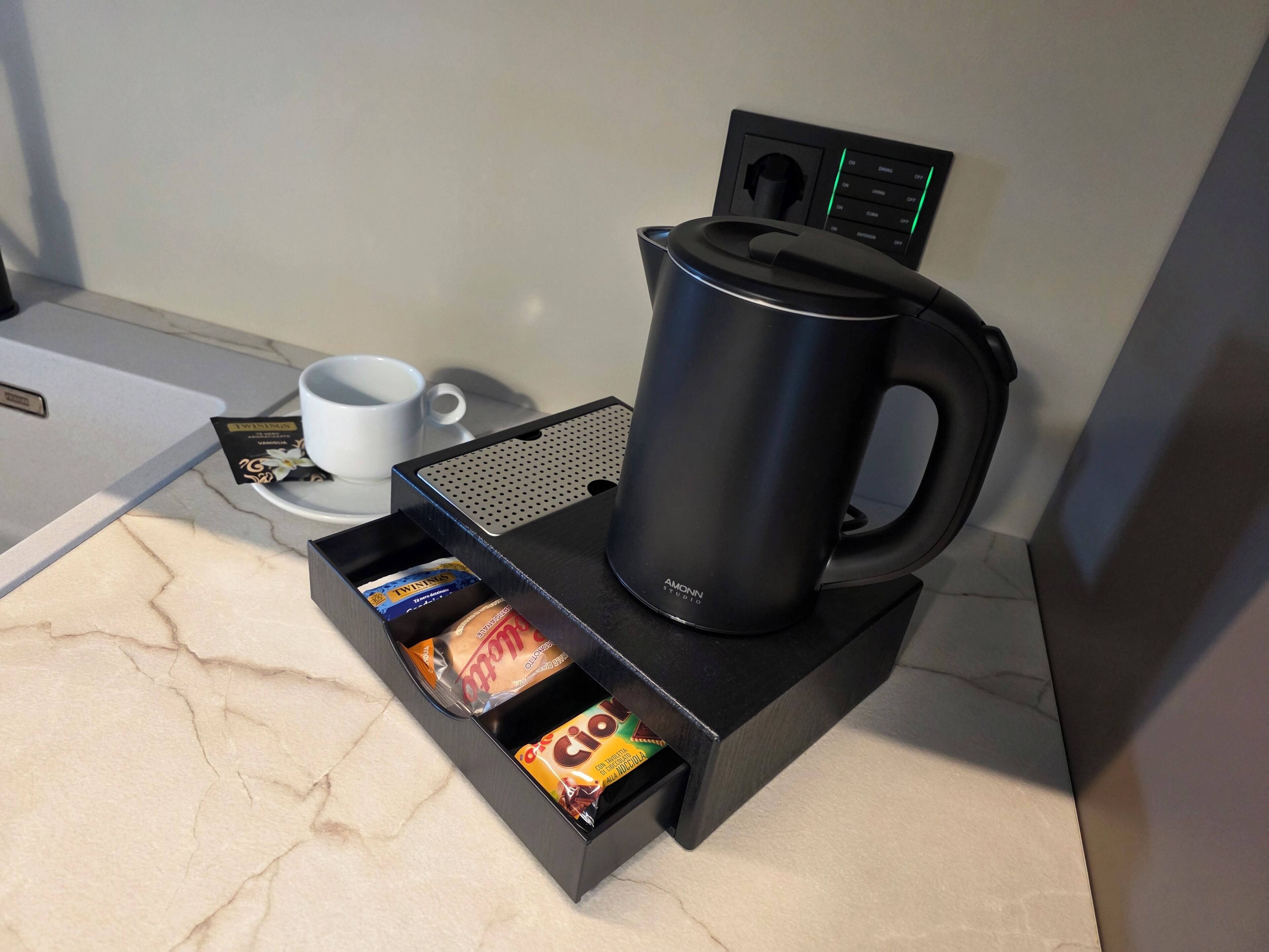 A coffee maker on a black stand with a coffee pod holder below it, a white cup, and a power strip with a control panel.