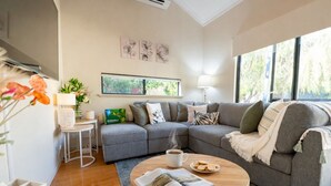 A modern living room with a grey sectional sofa, a coffee table with a magazine, a small side table with a lamp, and a window with a view of greenery. - "Serenity & Sea", Pool, Wi-Fi, Internet, Netfilix/Stan, 500m to Beach, Parking (Preston Beach)