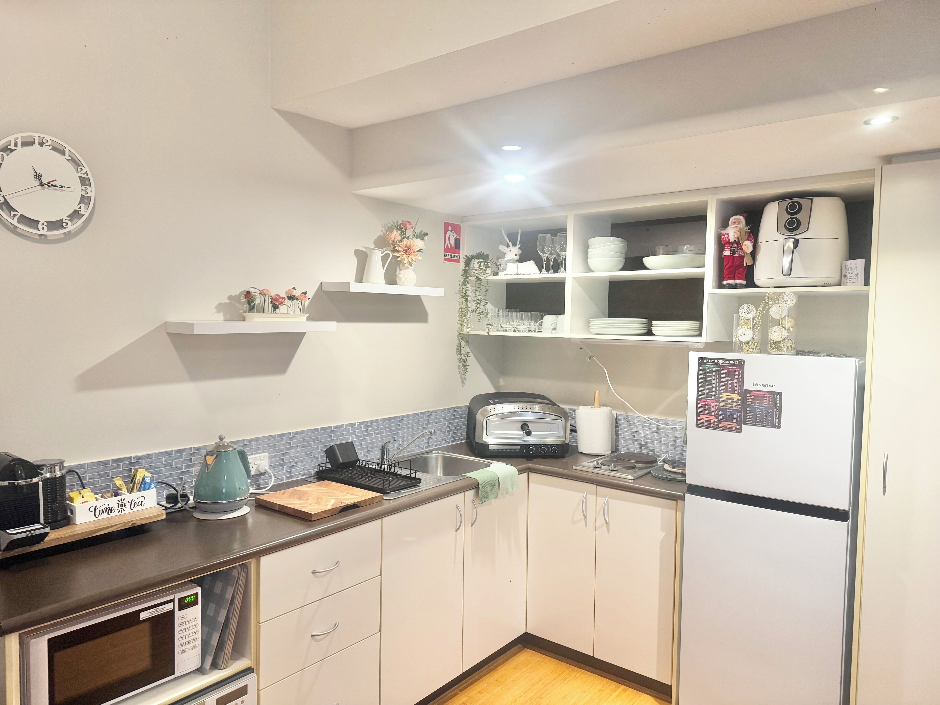 A modern kitchen with white cabinets, a microwave, a toaster, and a refrigerator. There are shelves with decorative items and a clock on the wall.
