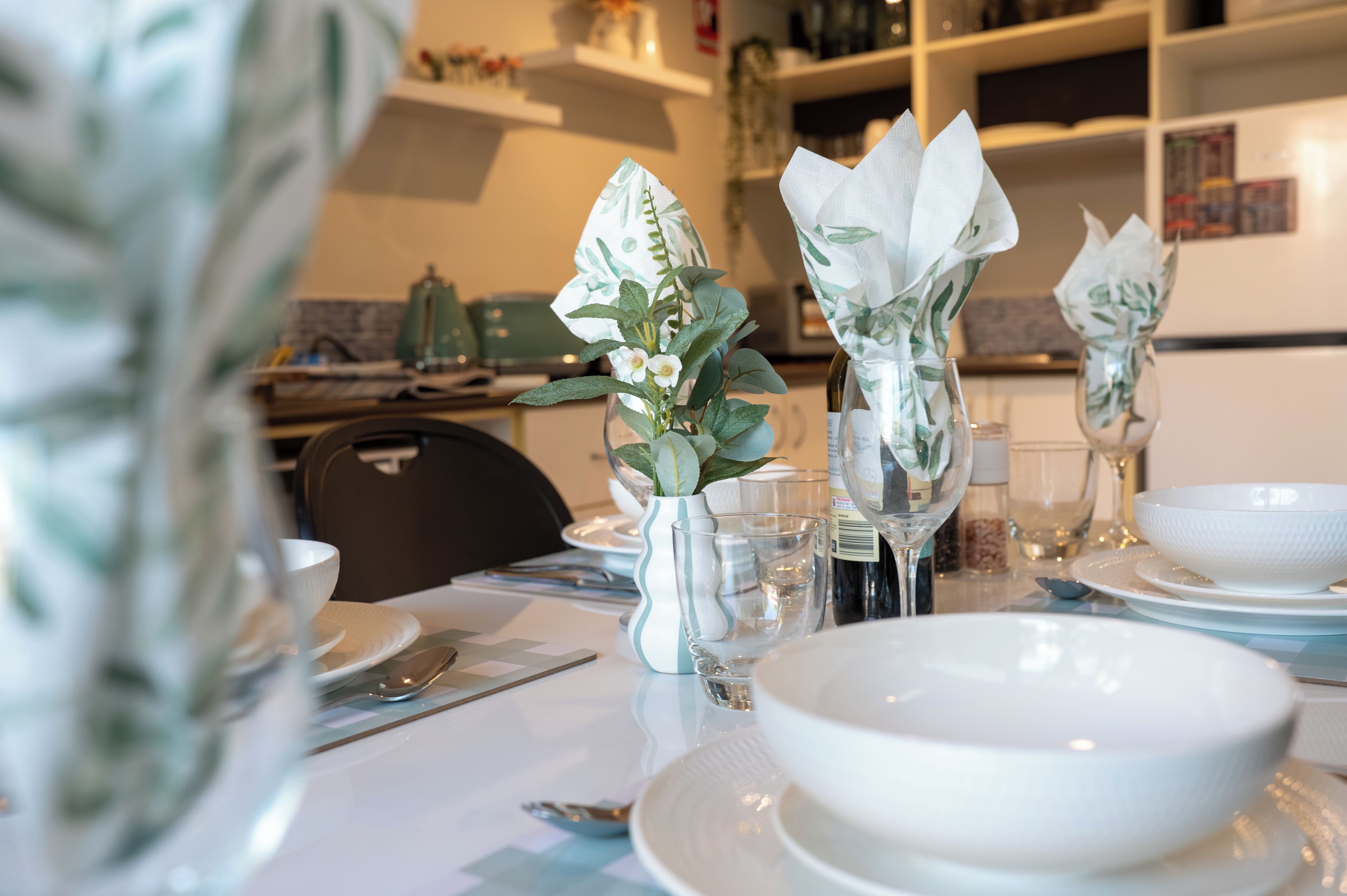 Taula de dinar coberta amb plats, bols i recipients de vidre blancs, decorada amb flors de paper i un ram d'herbes.