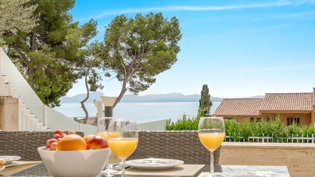 A table set with breakfast items, including a bowl of fruit and glasses of orange juice, overlooking a scenic view of a lake and mountains. - Miramar Apartment with sea views (Alcúdia)