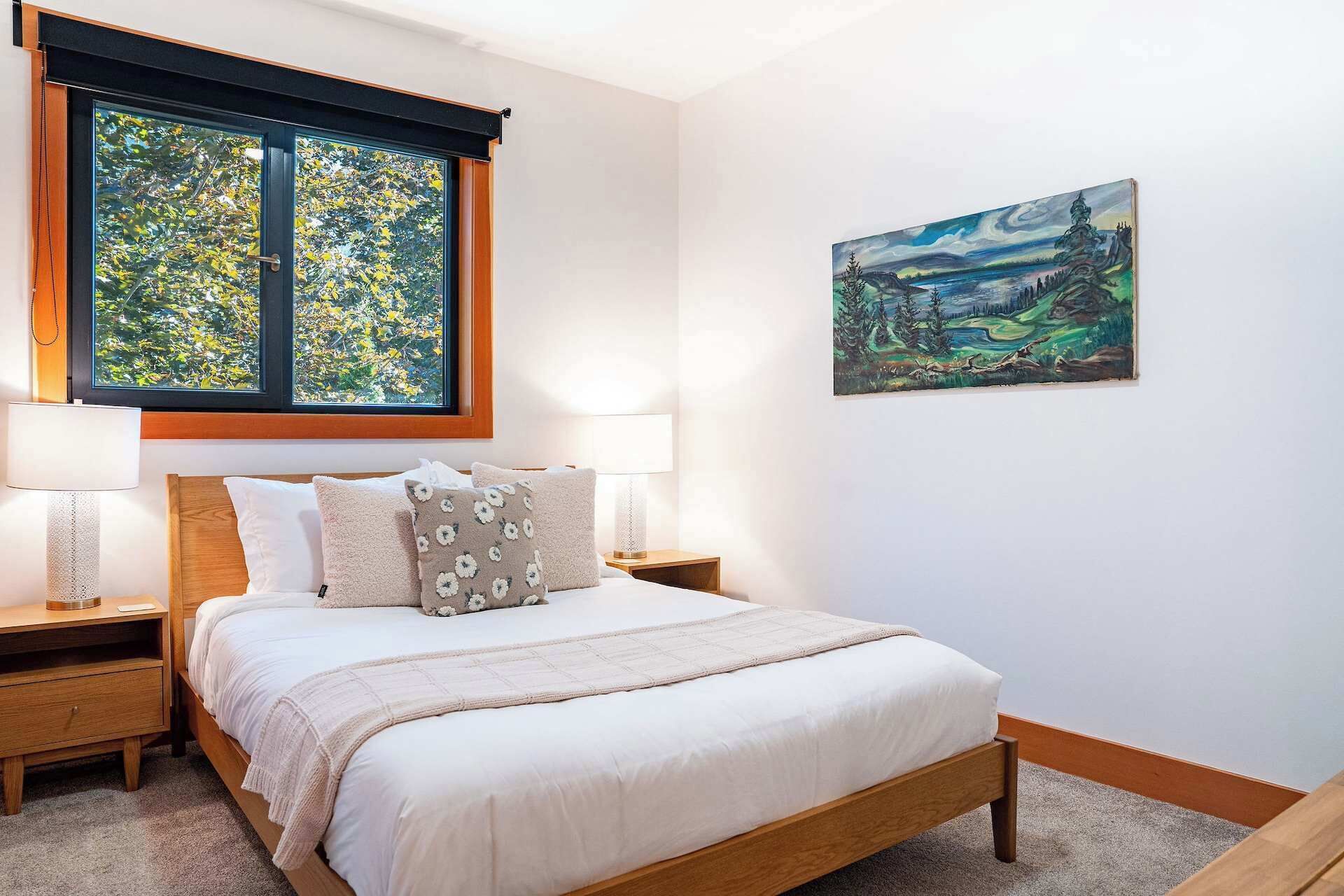 A bedroom with a bed, two nightstands, a window with a view of trees, and a painting on the wall.
