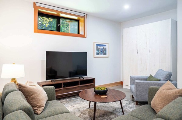 A living room with a flat-screen TV, a sofa, a coffee table, and a window with a view of trees.
