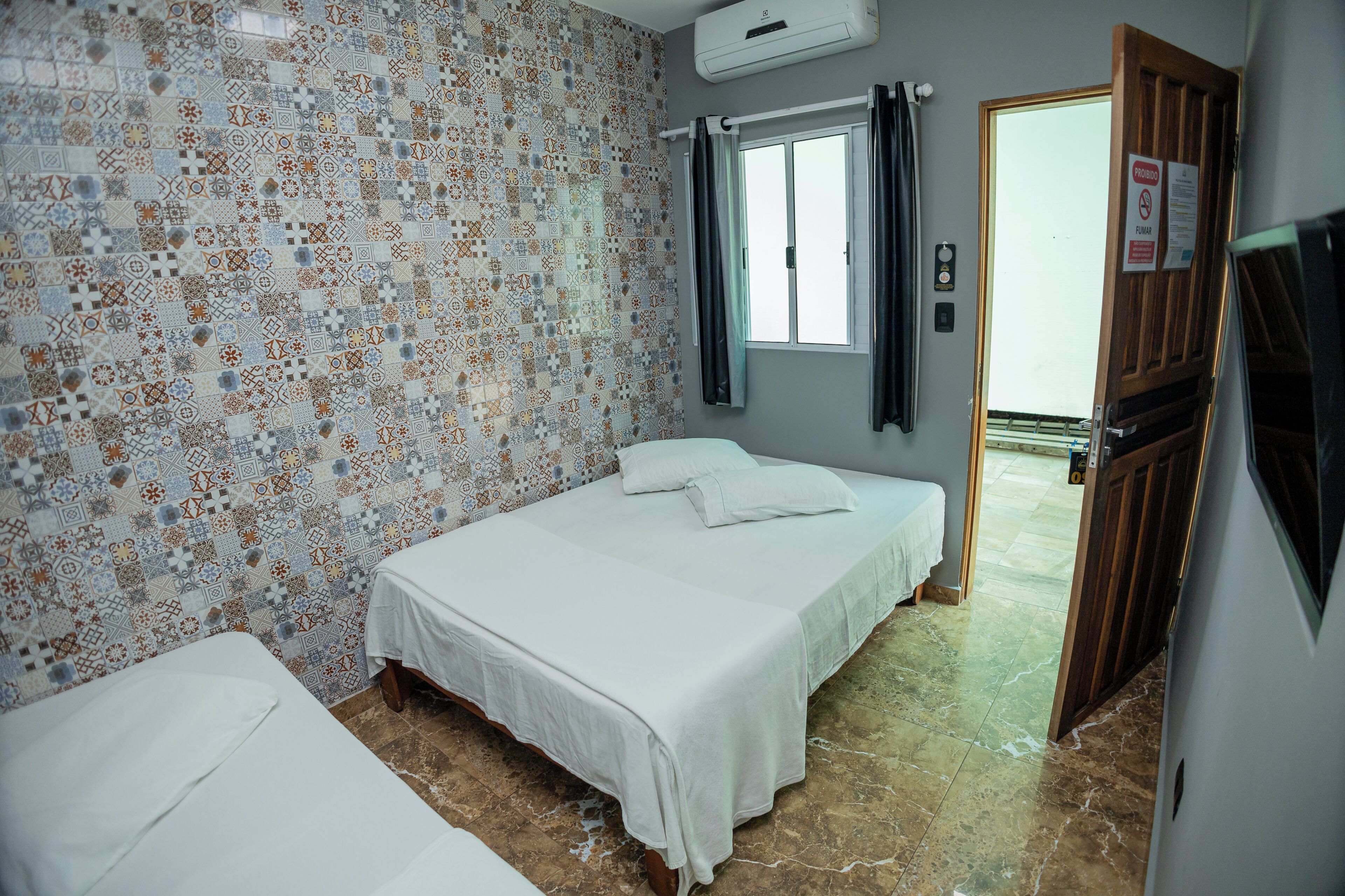 A hotel room with a patterned tile wall, a bed with white bedding, a window with curtains, and a door with a sign.