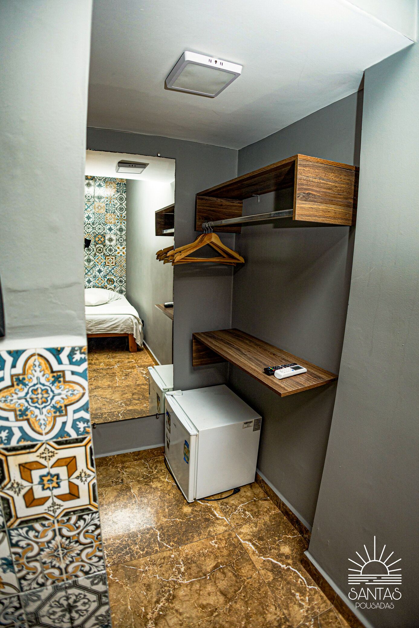 Quarto de hotel compacto com cama, pequeno frigorífico, prateleira de madeira e espaço para pendurar roupas.