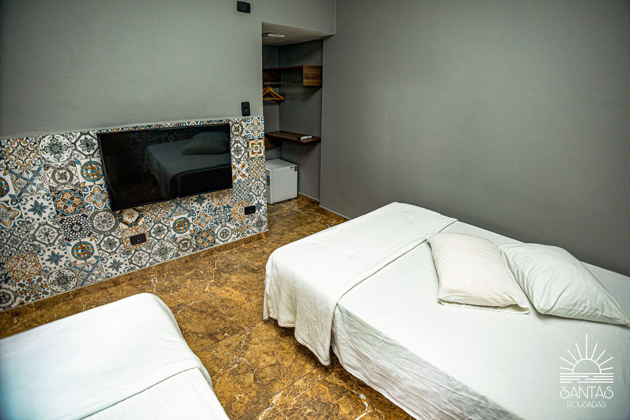 A hotel room with two beds, a television mounted on a patterned wall, a small refrigerator, and a wooden shelf with a hanging garment.