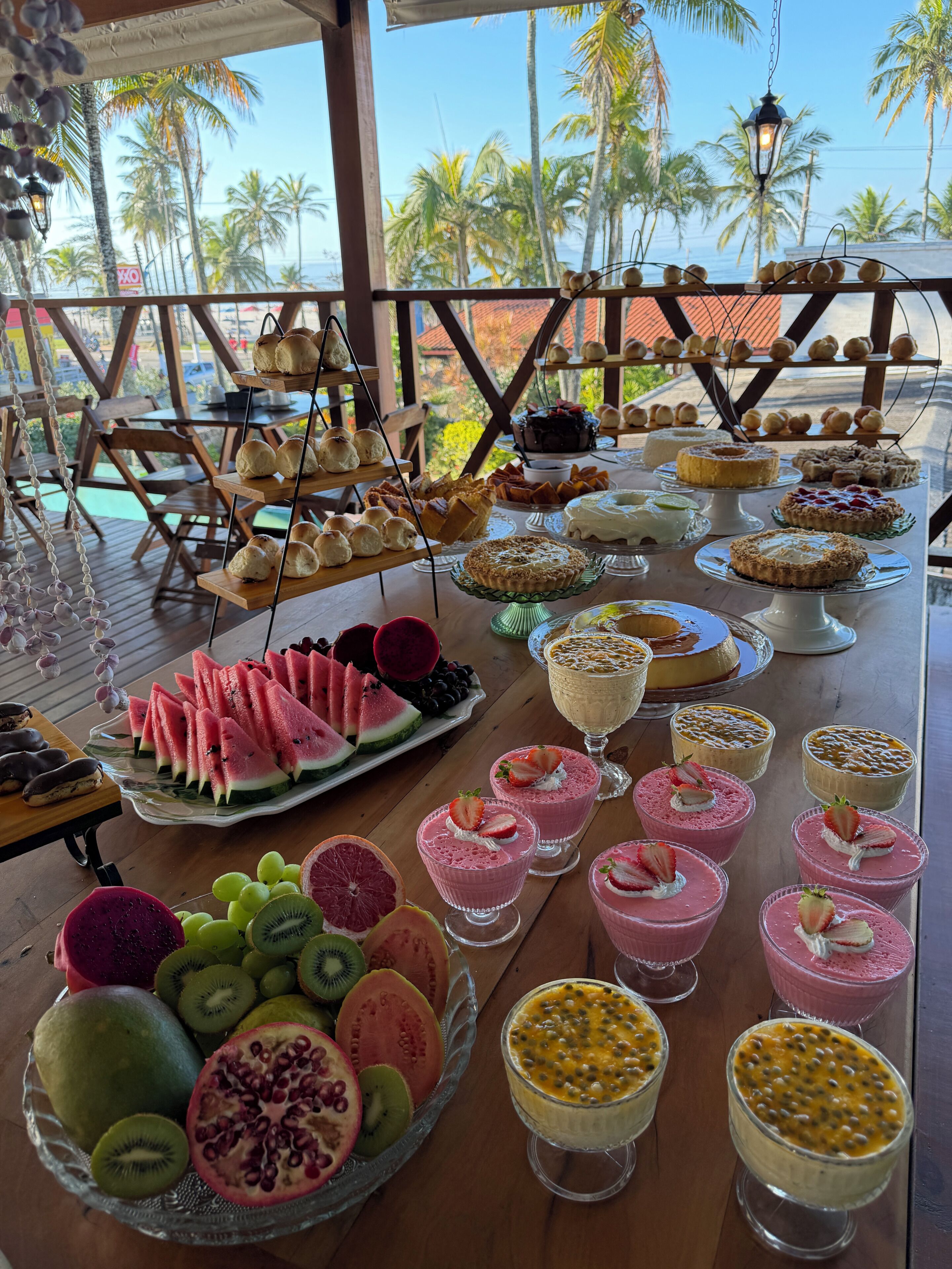 Une table de buffet proposant divers desserts et fruits, avec vue sur une plage bordée de palmiers.