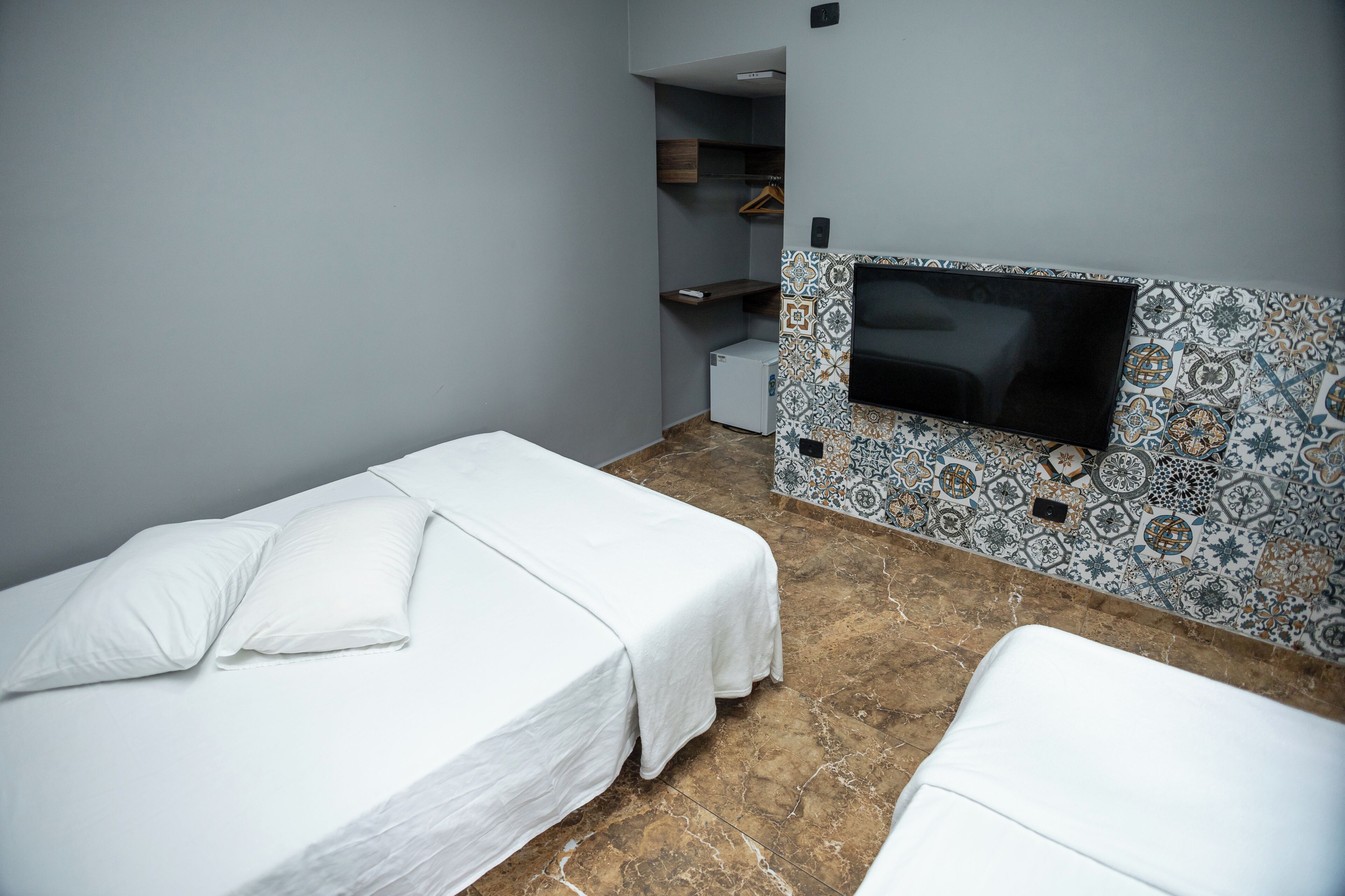 A hotel room with two single beds, a television mounted on a patterned wall, and a small refrigerator.