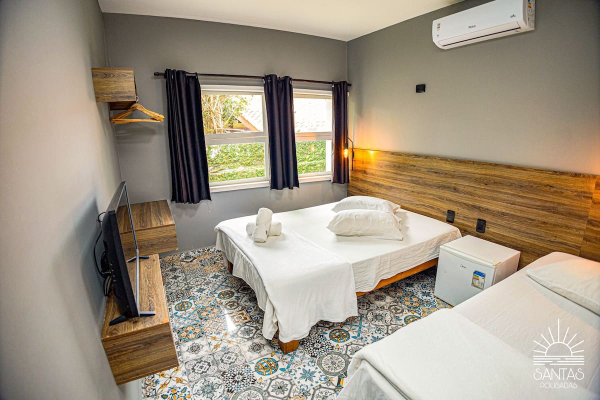 A hotel room with a bed, a small refrigerator, a wooden side table, a TV, and a window with curtains.