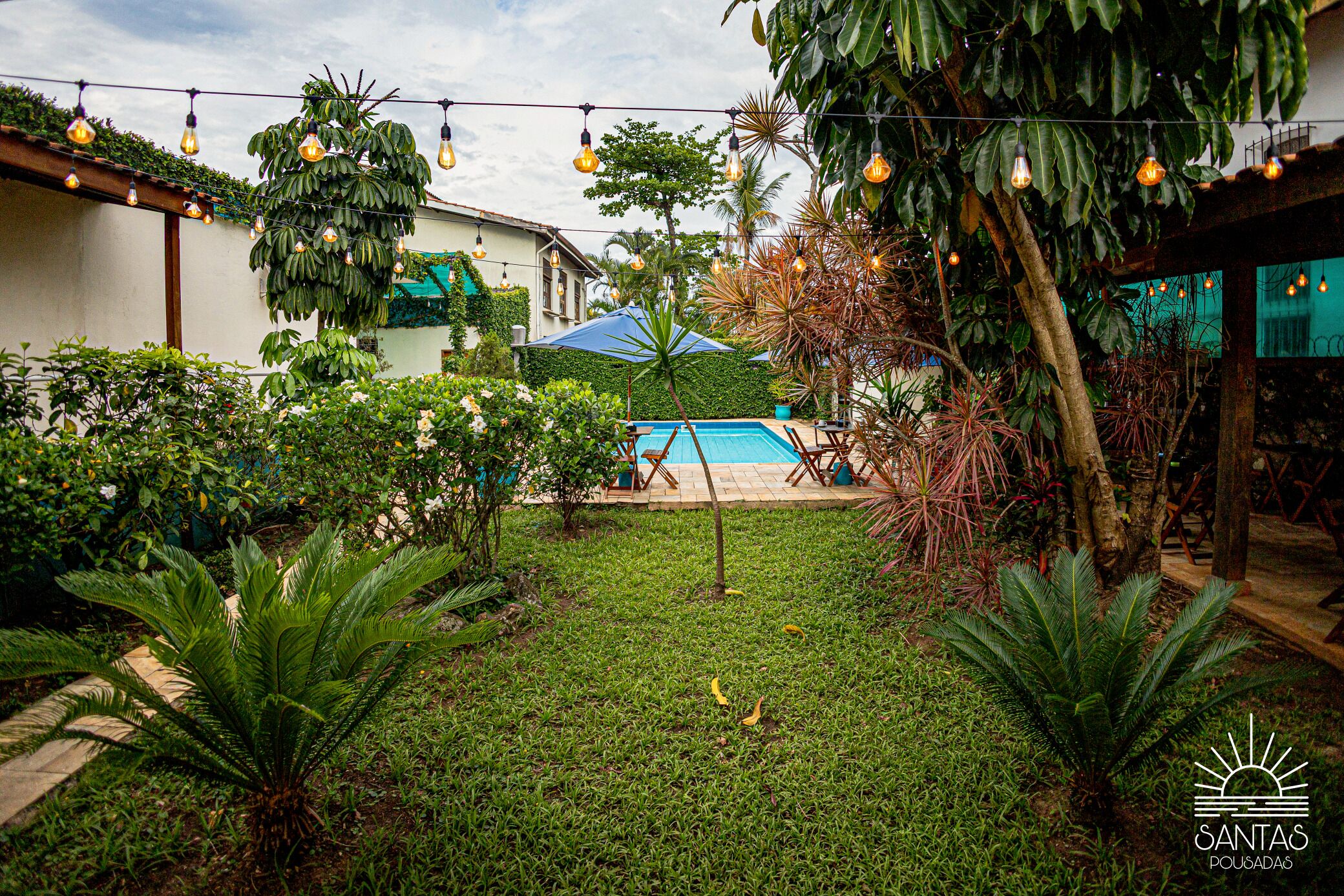 Un jardin avec une piscine, entouré d’une végétation luxuriante et illuminé de guirlandes lumineuses.