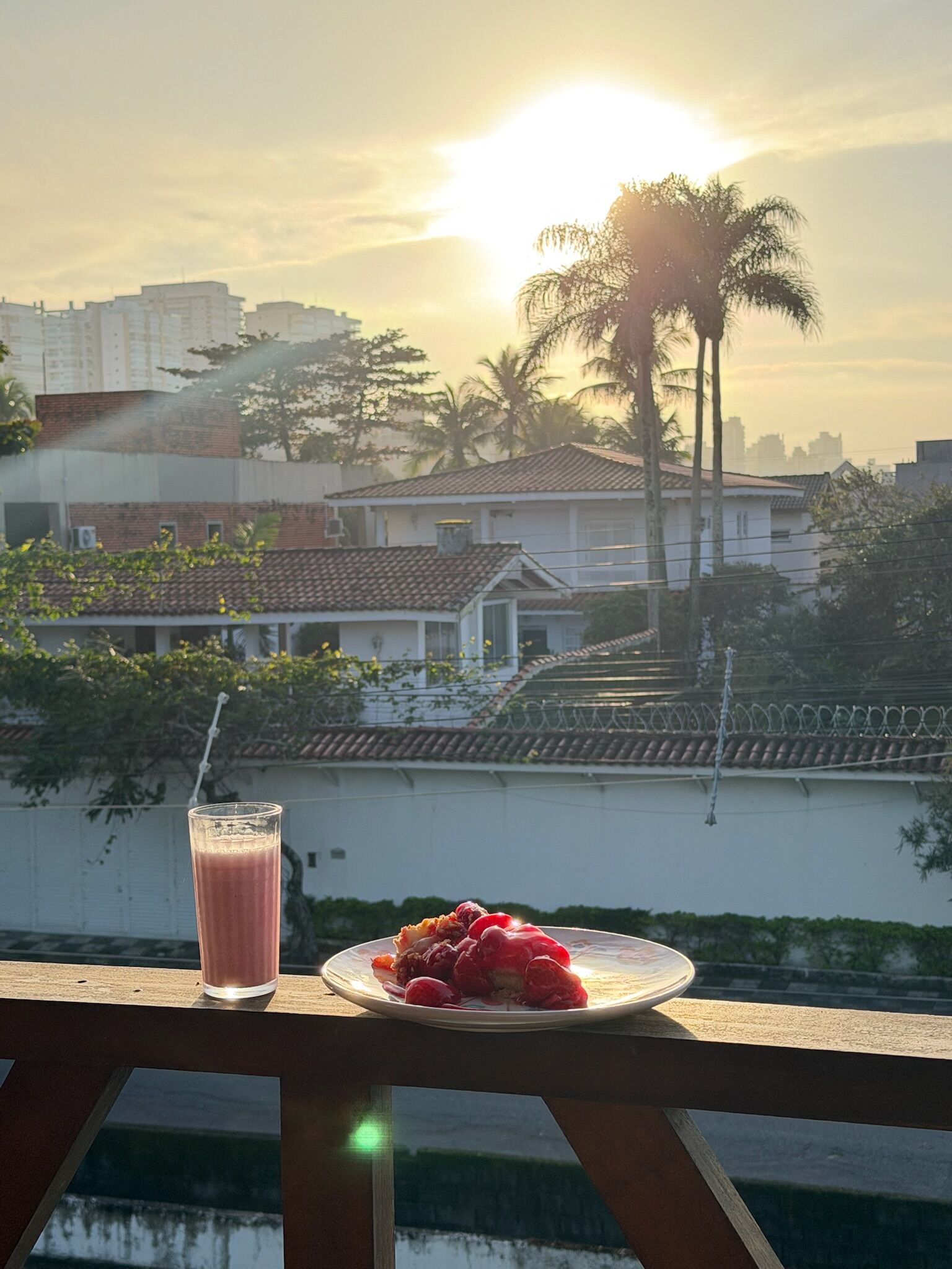 A balcony with a glass of beverage and a plate of fruit, overlooking a residential area with buildings and palm trees.