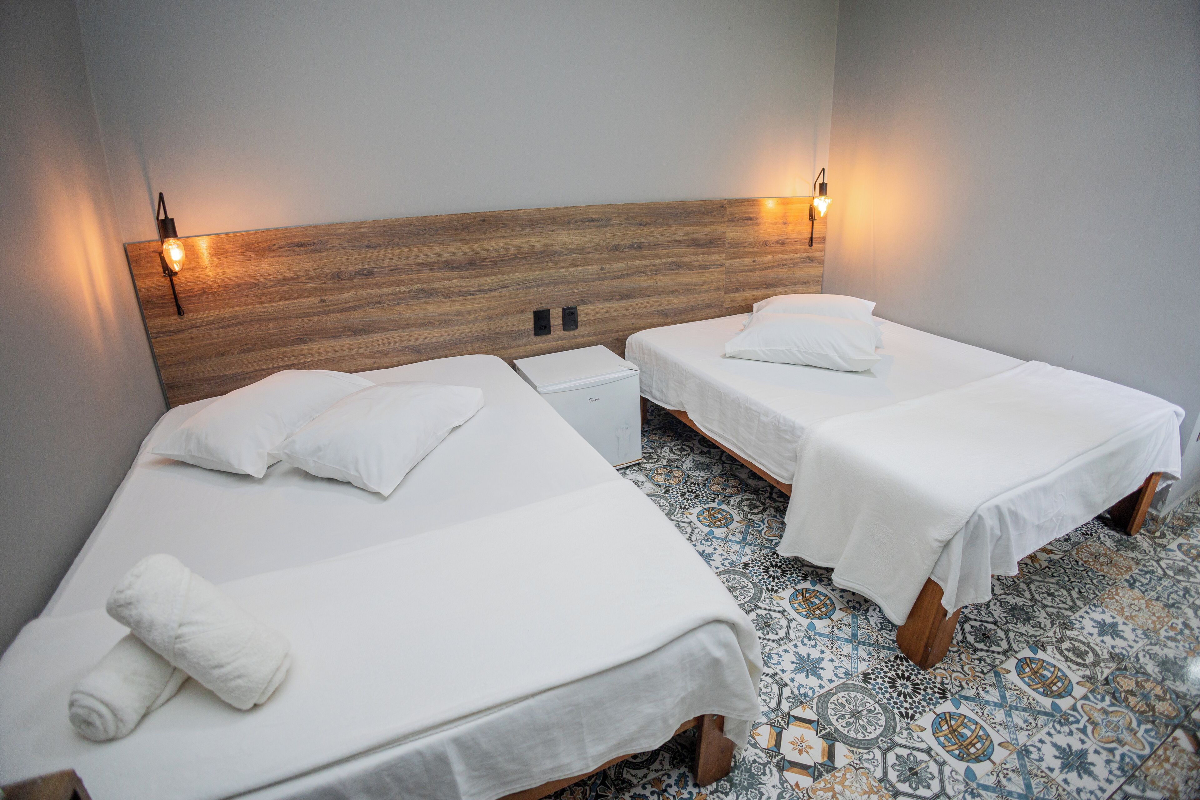 A hotel room with two single beds, a wooden headboard, bedside lamps, and patterned floor tiles.