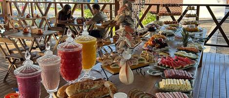 A buffet table with various food items and drinks, located under a wooden structure with a view of the beach and palm trees.