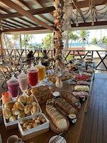 A buffet table with various food items and drinks, located under a wooden structure with a view of the beach and palm trees.
