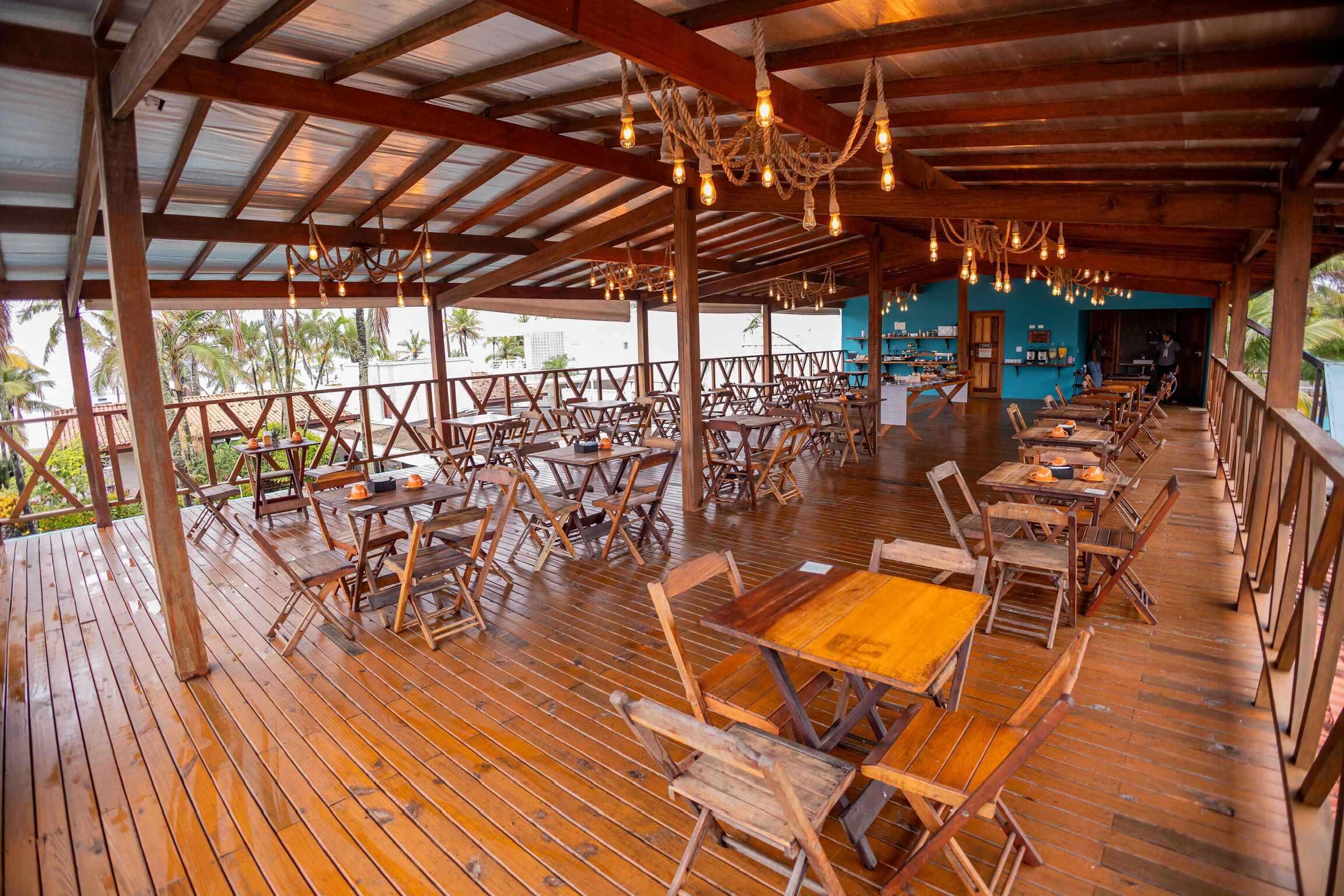 An outdoor dining area with wooden tables and chairs, a covered roof, and hanging light fixtures.