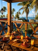 A wooden table set with breakfast items and orange juice, overlooking a beach with palm trees and the ocean.