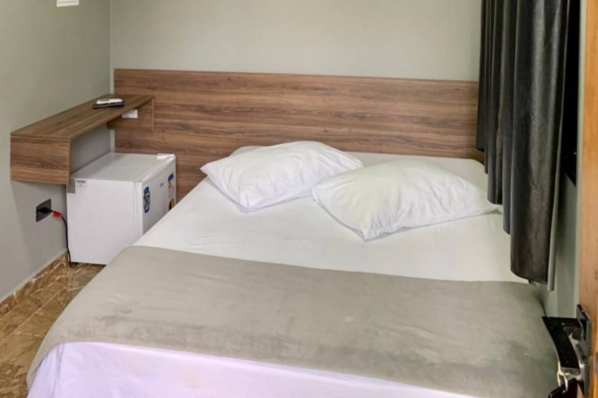 A hotel room with a bed, a wooden headboard, a small wooden shelf, a heater, and a black curtain.