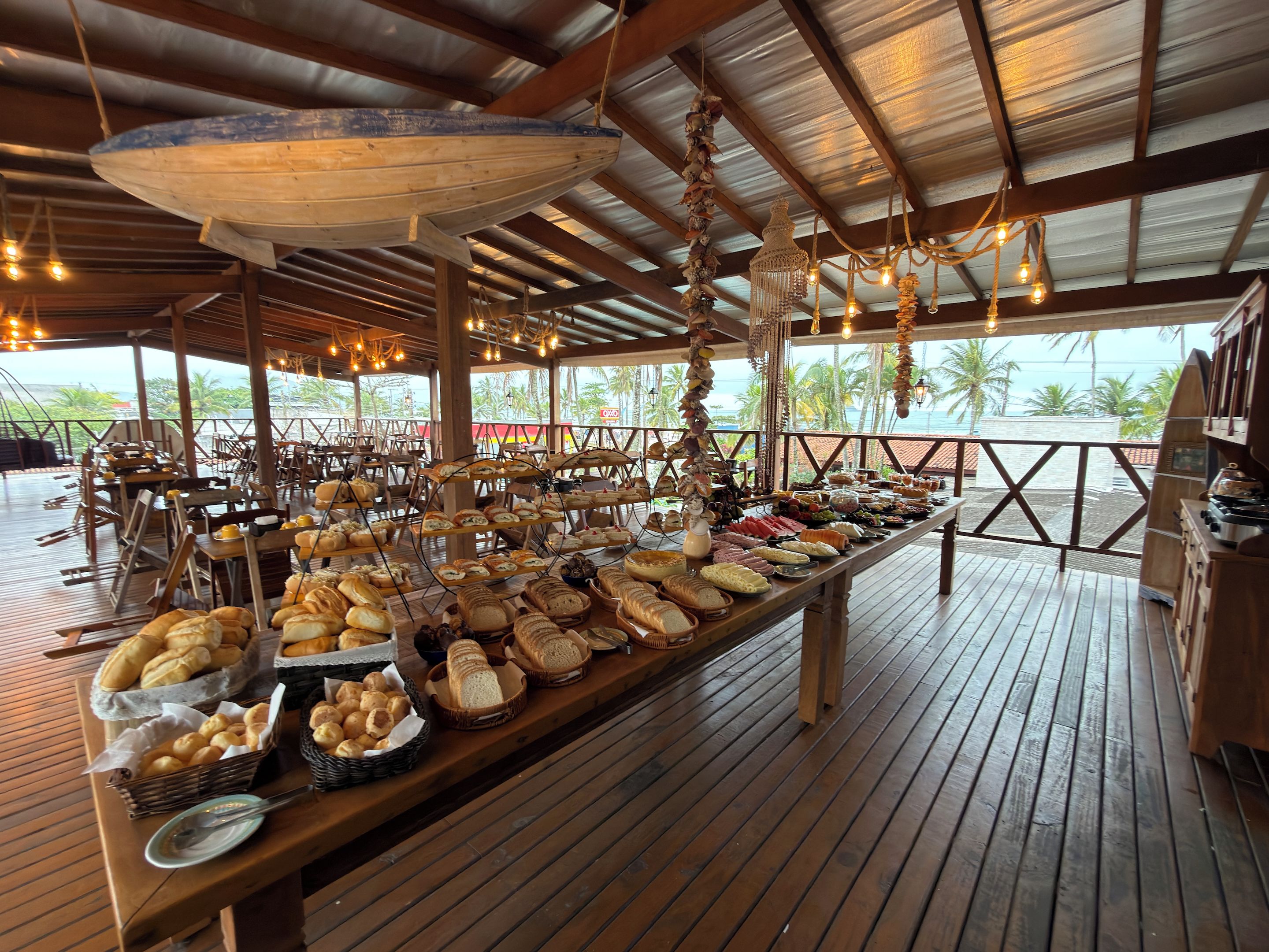 Un buffet proposant une variété de plats, disposé sur une table en bois, sous une zone abritée agrémentée de guirlandes lumineuses, avec vue sur l’océan.
