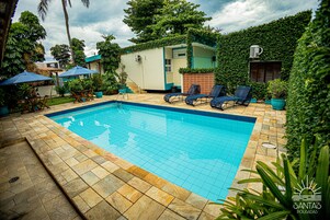 Piscina com piso em azulejos, cercada por espreguiçadeiras e guarda-sóis, e um pequeno edifício com telhado verde.