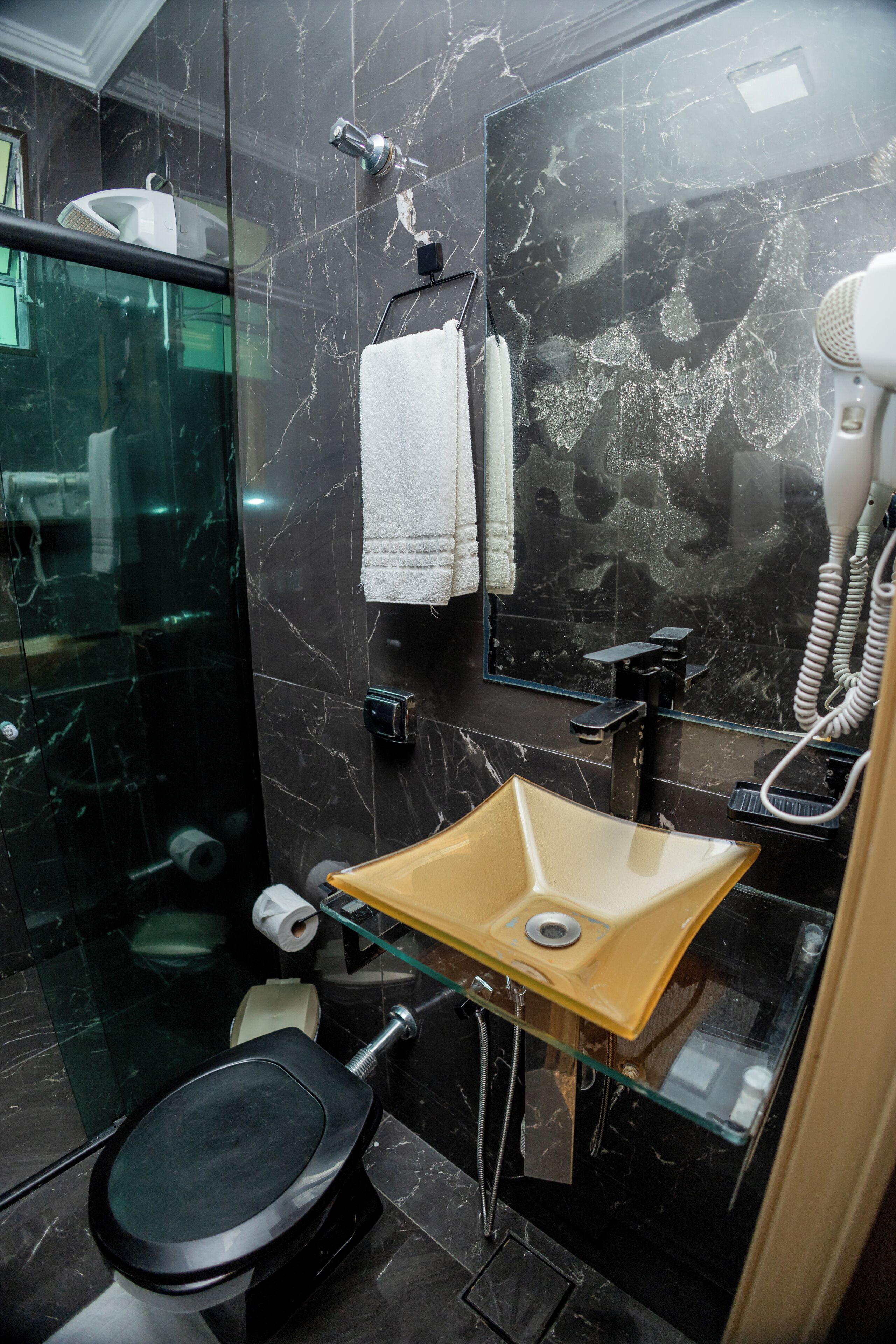 Casa de banho moderna com chuveiro em cabine de vidro, sanita preta, lavatório de vidro e secador de cabelo.