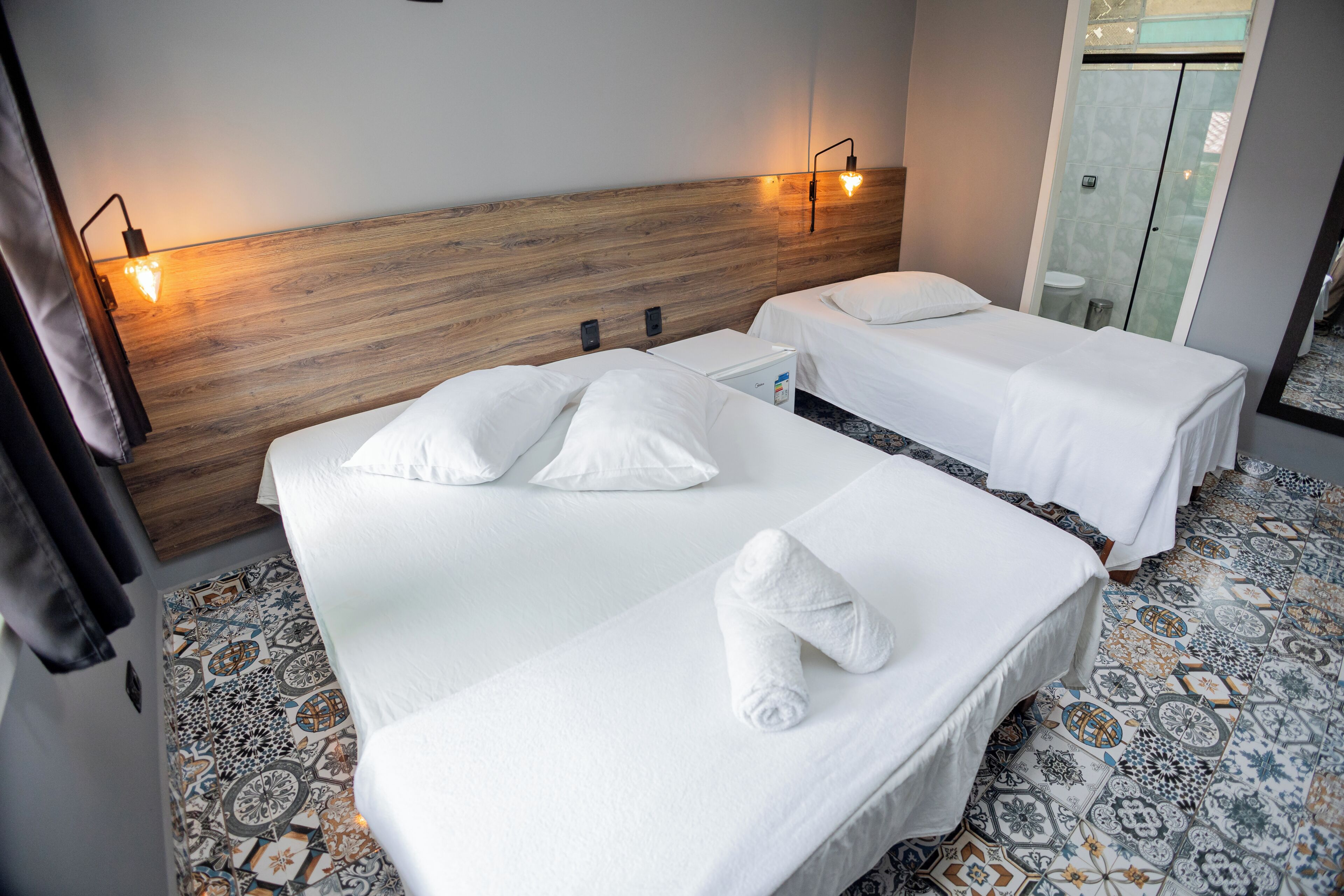 Une chambre d’hôtel avec deux lits, une tête de lit en bois, un minibar et une salle de bain avec une douche en verre.