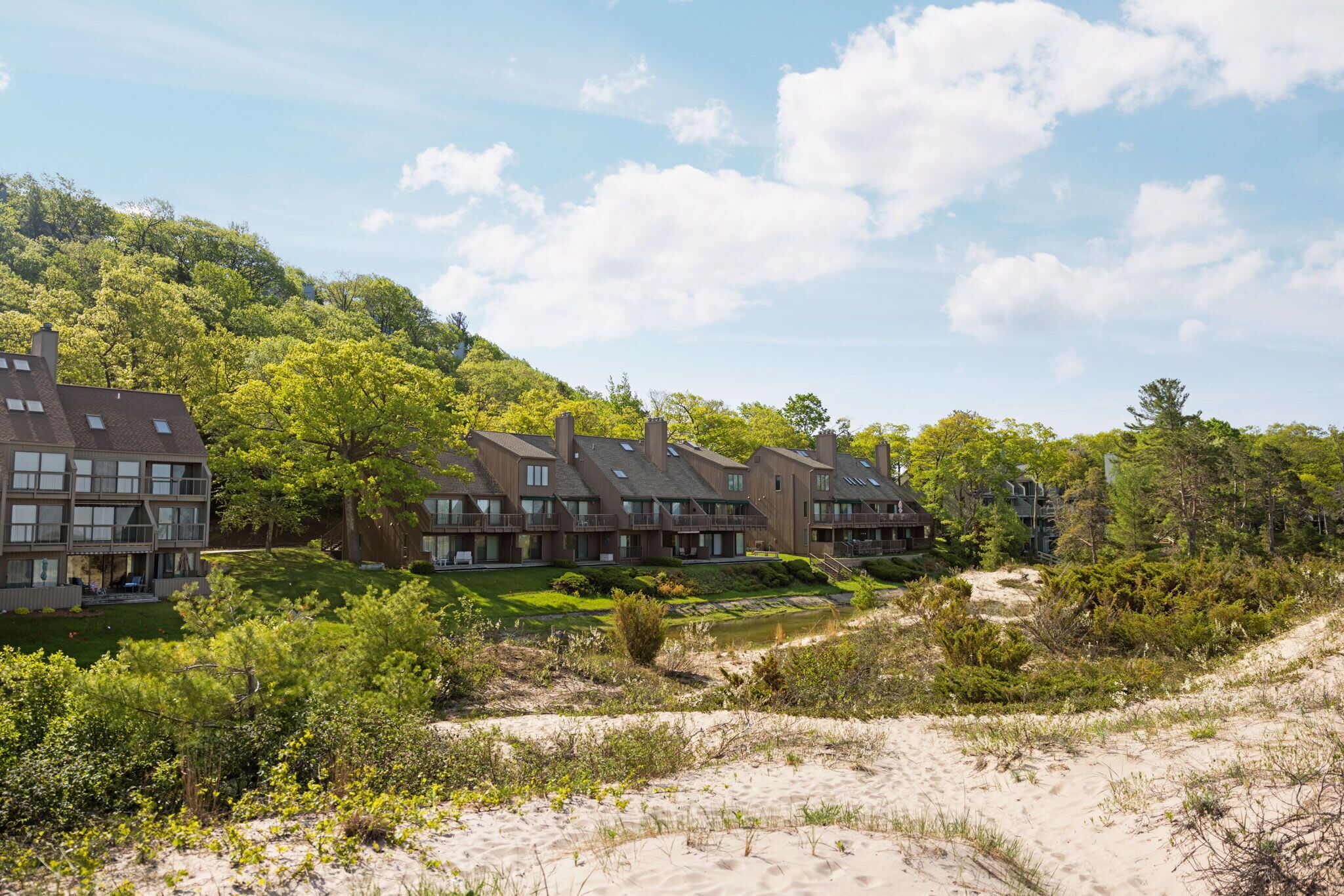 A row of modern multi-story buildings with balconies, set against a backdrop of lush greenery and a sandy landscape.