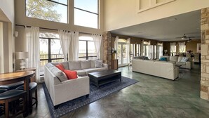 Amplia sala de estar con un sofá seccional grande, un sofá más pequeño y una mesa de centro. Hay un comedor con mesa y sillas. La habitación tiene ventanas grandes y vistas al exterior.