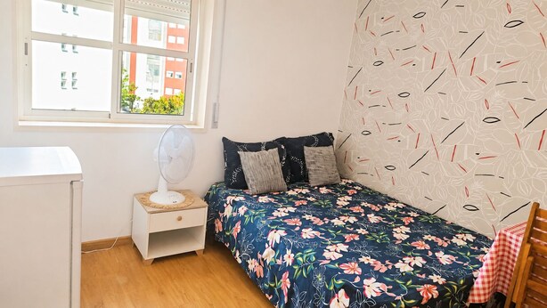 A bedroom with a bed, a bedside table with a fan, a window, and a patterned wall. - Room-charming Lisbon Apartment in Quiet (Lisbon)